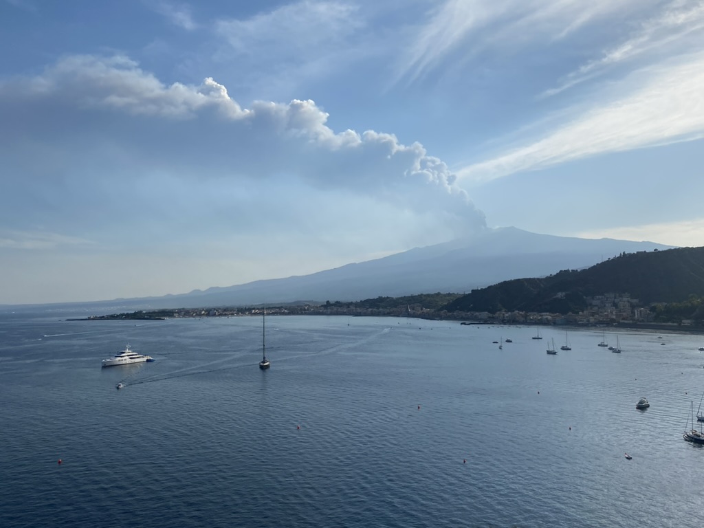 View of Mount Etna emitting smoke, with boats on the calm sea in the foreground.