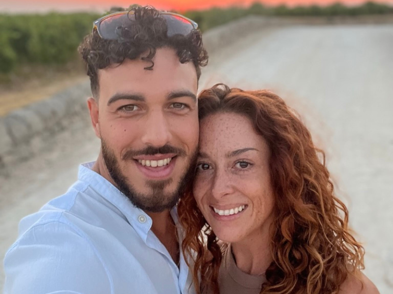 Alessio and Monika smiling together at sunset in Sicily, with a warm, orange-pink sky and vineyard in the background.